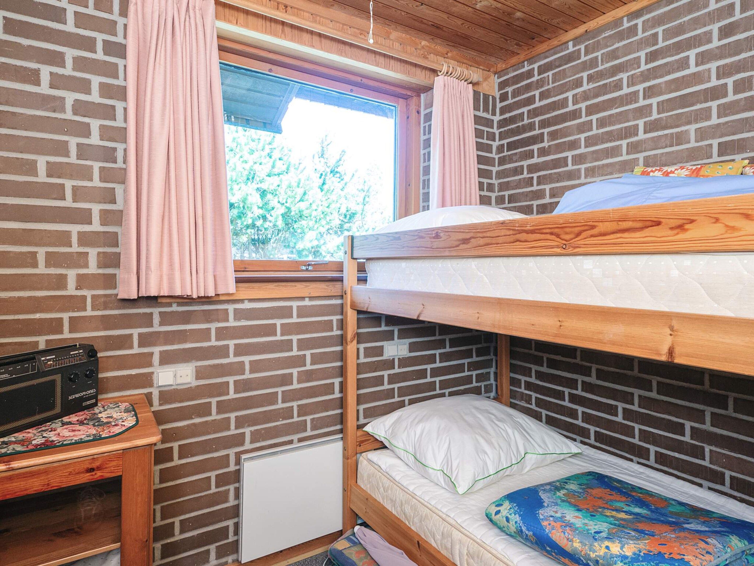 Habitación con literas, pared de ladrillo, ventana con cortinas, escritorio de madera con radio y un colchón con almohada colorida.