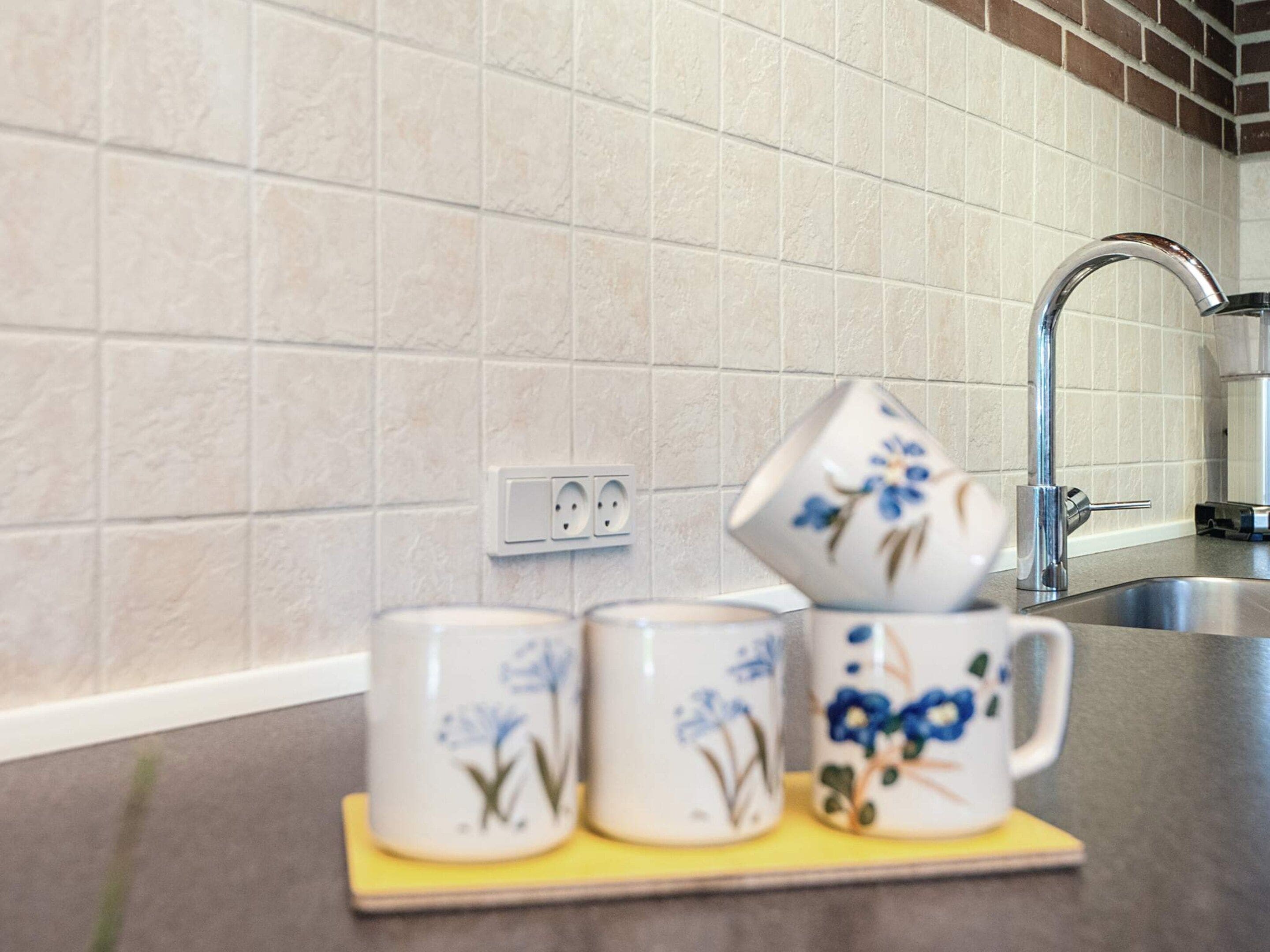 A kitchen with a sink, a faucet, and a countertop with three ceramic mugs on a coaster.