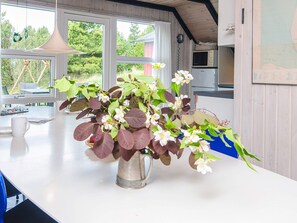 A kitchen with a white countertop, a metal pitcher with flowers, and a window with curtains.