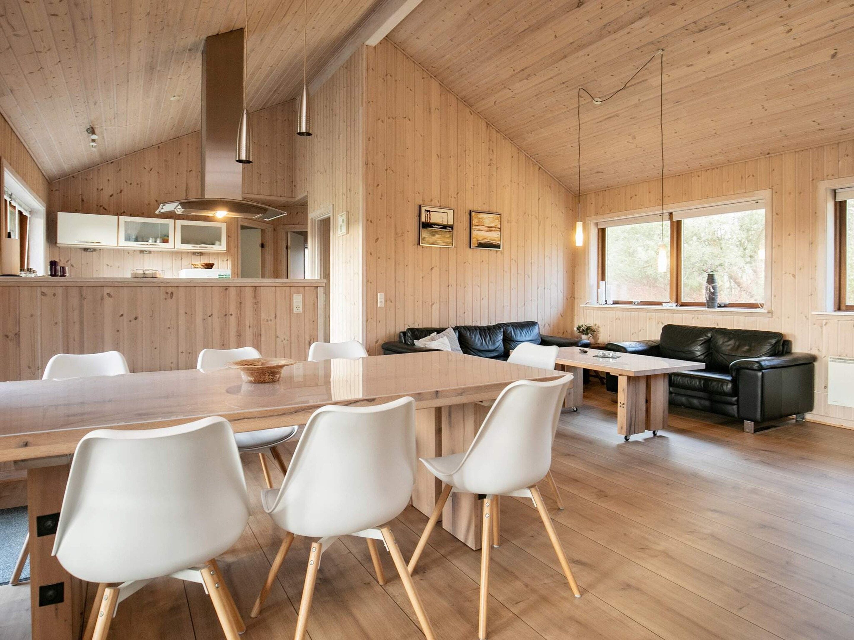 A spacious room with a wooden ceiling, a dining table with chairs, a black leather sofa, and a window with a view of trees.