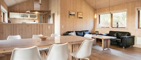 A spacious room with a wooden ceiling, a dining table with chairs, a black leather sofa, and a window with a view of trees.