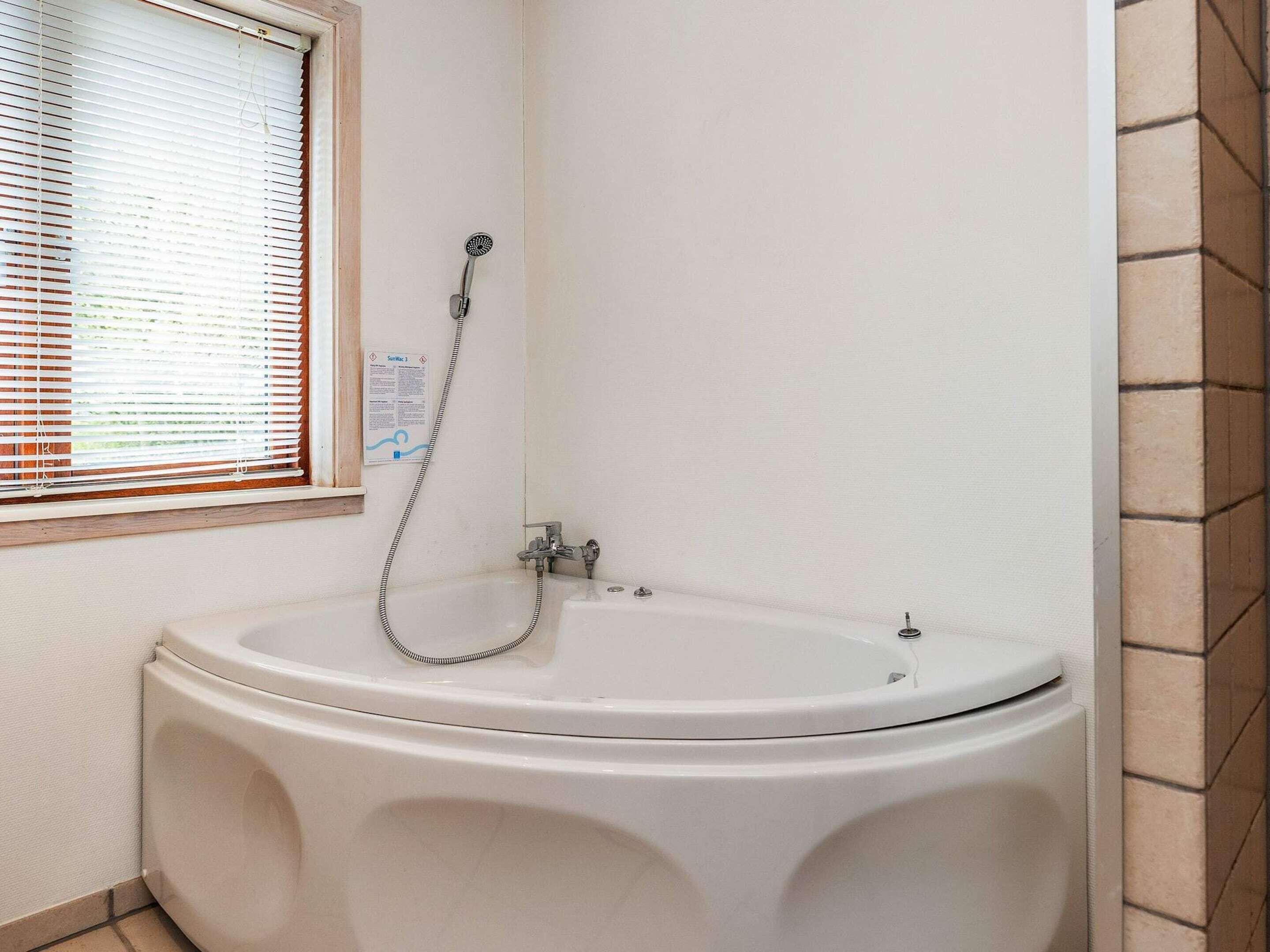 Une salle de bain avec une grande baignoire, une fenêtre avec des stores et une pomme de douche.