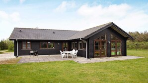 A black wooden house with a tiled roof, a patio with white chairs, and a grassy yard. - 4 Star Holiday Home in Lokken (Lokken)