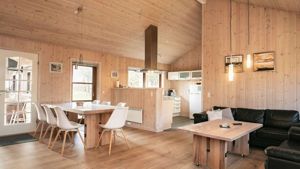 A spacious room with a wooden floor and walls, a dining area with a table and chairs, a black leather sofa, and a kitchen area with cabinets and a refrigerator. - 4 Star Holiday Home in Lokken (Lokken)