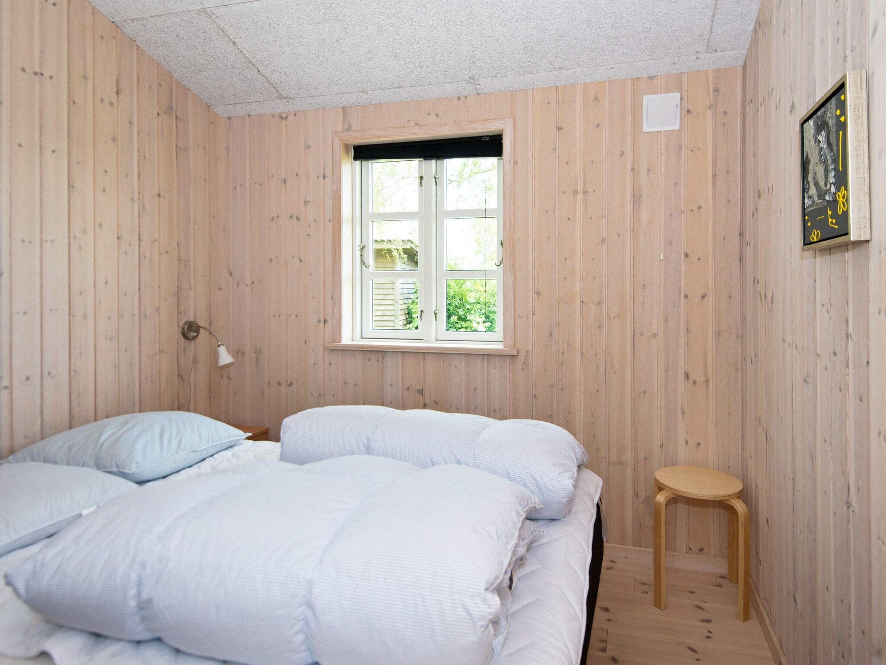 Ein Zimmer mit einem Bett, einem Fenster, einem Hocker und einer Wandleuchte.