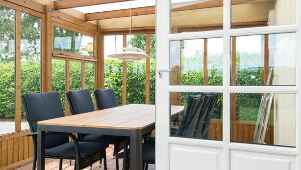 En overdækket terrasse med et træbord og stole, store vinduer og en hængende lampe.