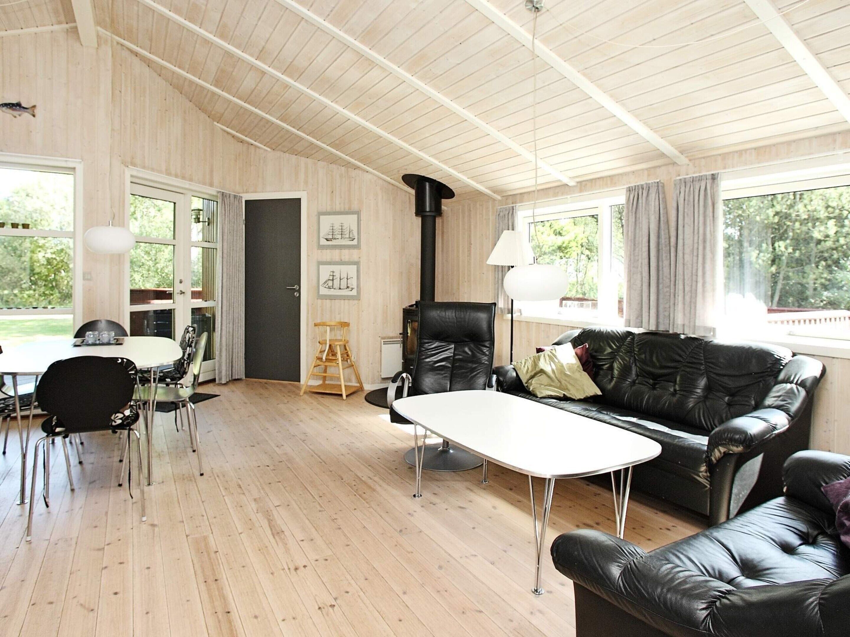 En rummelig stue med trægulv, en sort lædersofa, et hvidt bord og en sort læderlænestol. En dør fører ud til et udeområde, og en lampe er synlig på højre side.