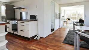 A modern kitchen with white cabinetry, a stainless steel range hood, and a built-in oven. There is a dining area with a white table and chairs, and a view of the outdoors through large windows. - Stunning Holiday Home in Hovedstaden With Terrace (Gilleleje)