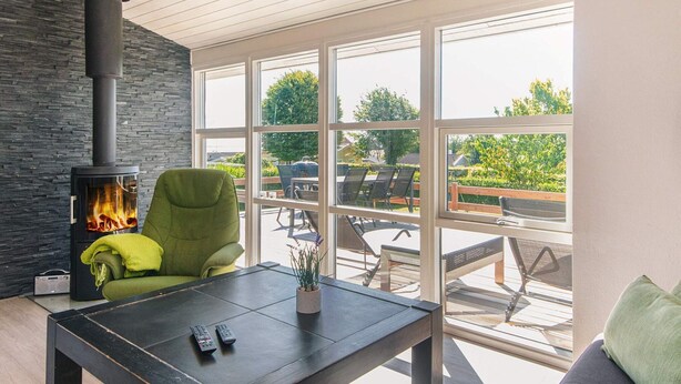 A modern living room with a stone wall, a black fireplace, a green armchair, a table with remotes, and large windows overlooking a deck. - Holiday Home in Hejls (Hejls)