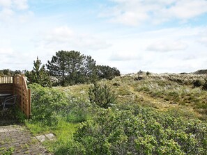 Una verja de madera da paso a un área pavimentada, con vistas a una colina cubierta de hierba y árboles.