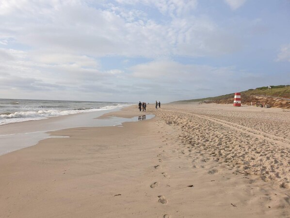 A sandy beach with footprints leading to the water, a red and white lifeguard tower, and a grassy hill in the background. - Cozy Holiday Home in Syddanmark near Beach (Ringkobing)