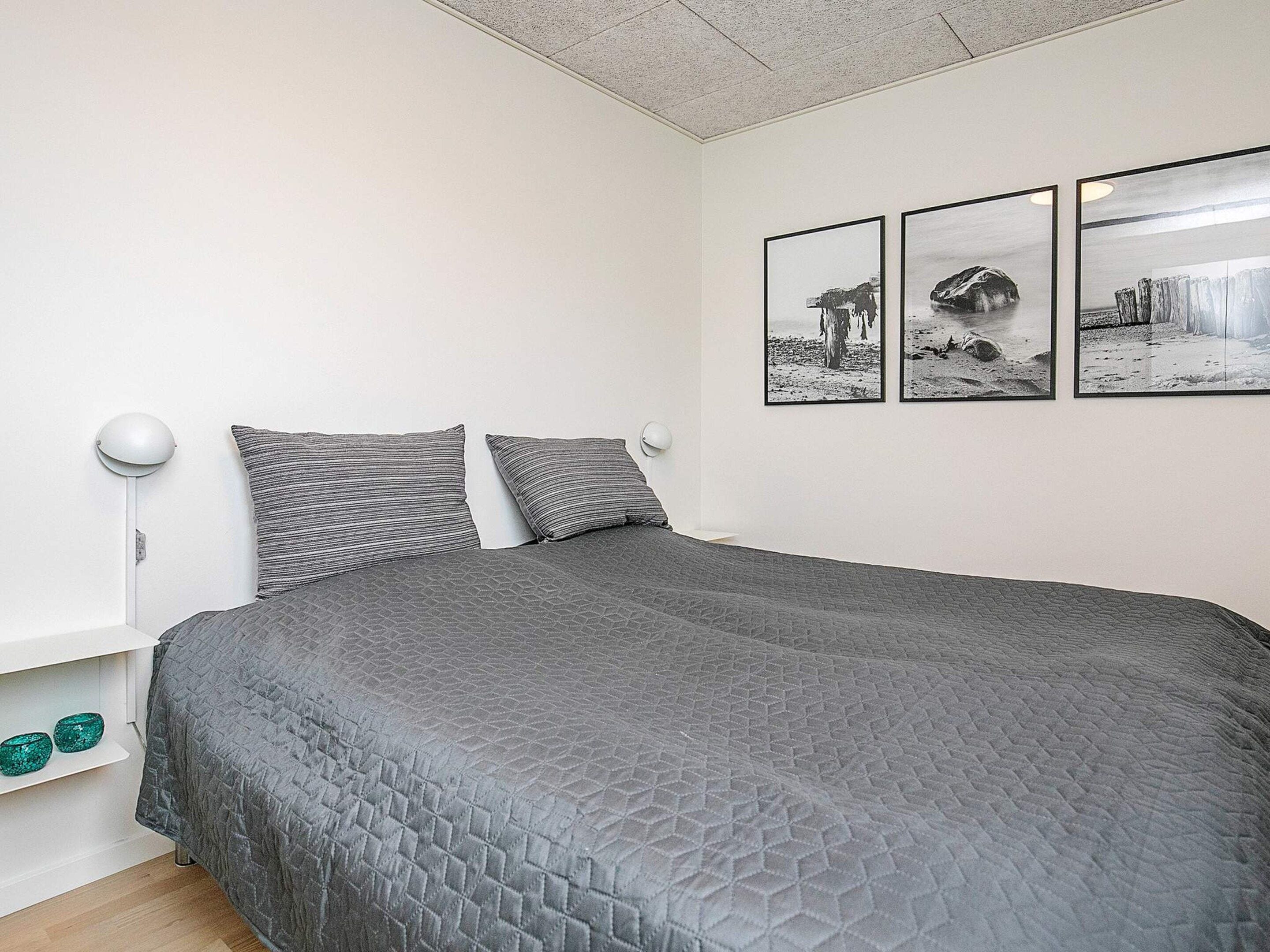A bedroom with a large bed, two bedside lamps, a shelf with decorative items, and three framed pictures on the wall.