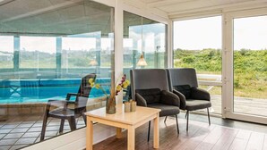 A modern room with a large window overlooking a swimming pool, a wooden table, and two grey armchairs. - 4 Star Holiday Home in Ringkobing (Ringkobing)