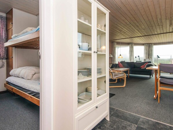A bunk bed with a cabinet containing dishes and glasses, a sofa, a small table, and a view through large windows.