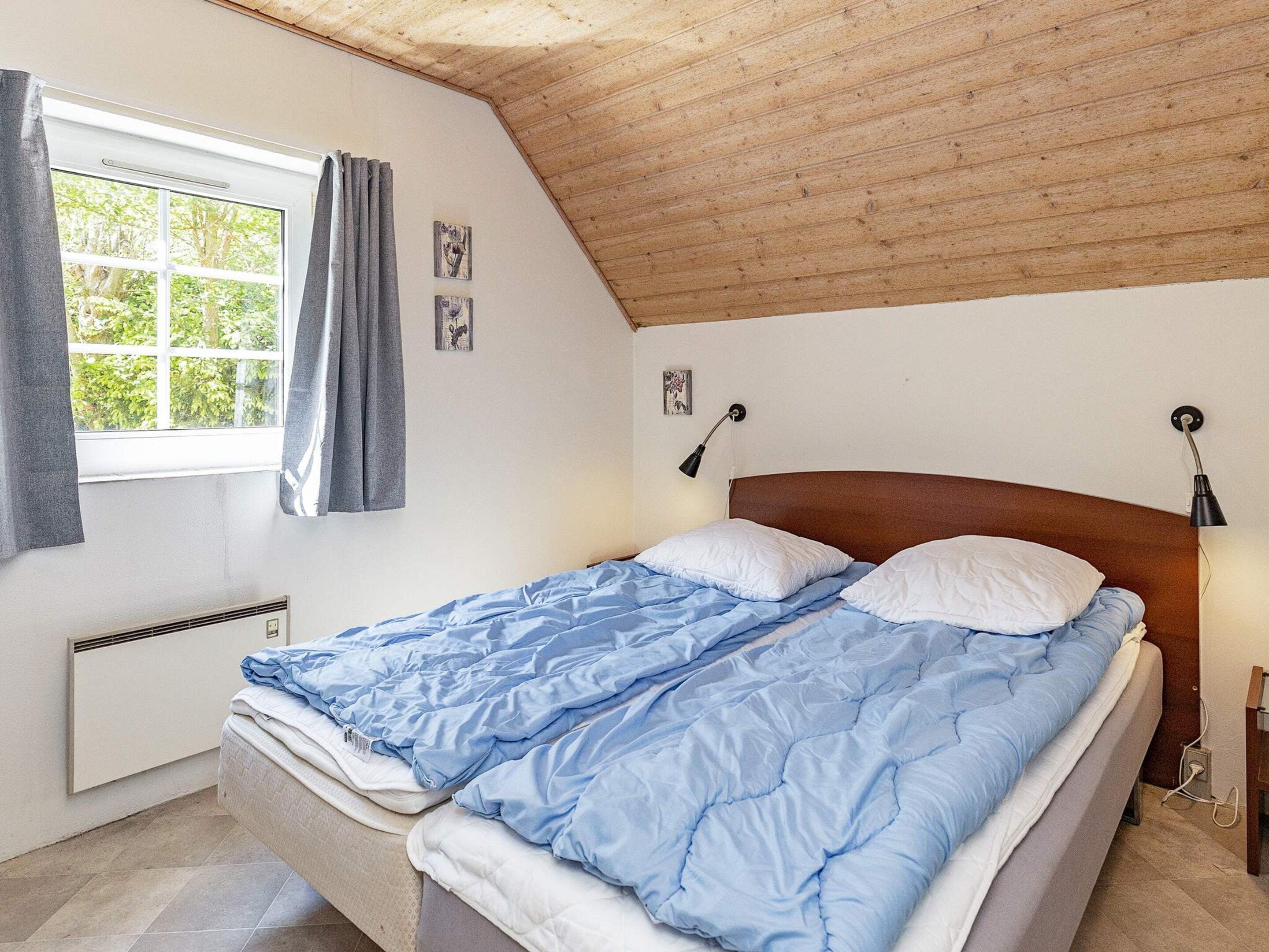 Une chambre avec un plafond en pente, un lit avec une literie bleue et blanche, une fenêtre avec des rideaux et des lampes fixées au mur.
