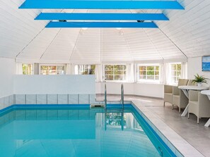 An indoor swimming pool with clear blue water, a tiled floor, and a ceiling with blue beams. There are windows allowing natural light and a seating area with chairs and a table. - 4 Star Holiday Home in Vaeggerlose (Vaeggerlose)