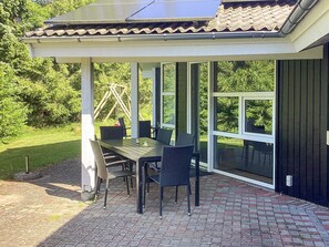 A covered patio with a table and chairs, large windows, and a view of greenery. - 4 Star Holiday Home in Vaeggerlose (Vaeggerlose)