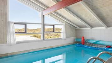 A swimming pool with clear blue water, a red lifebuoy, and a view of the beach through large windows.