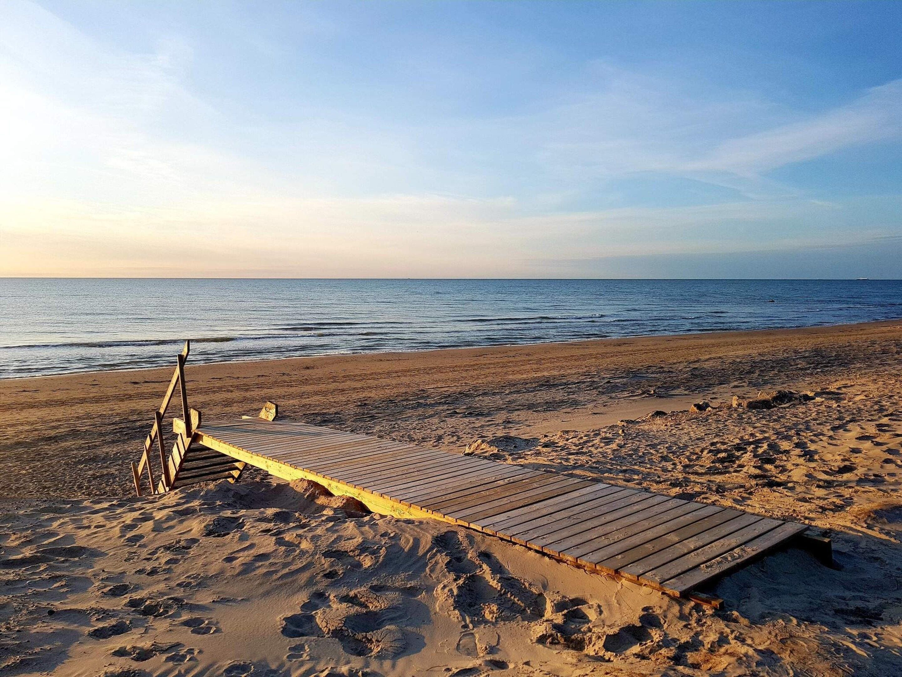 A beach with a wooden boardwalk leading to the water, footprints in the sand, and a clear sky.