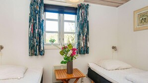 A room with two beds, a small wooden table with a vase of flowers, and a window with curtains. - 6 Person Holiday Home in Hvide Sande-by Traum (Hvide Sande)