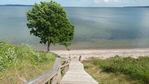 Una escalera de madera que lleva a una playa, con un árbol en primer plano y un mar tranquilo al fondo.