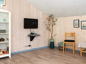 A room with a wooden wall, a mounted flat-screen TV, a small shelf with electronic devices, a vase with a branch, a wicker basket, and a wooden chair.