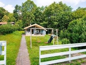 A wooden swing set and a covered seating area with a table and chairs, surrounded by lush greenery and a brick pathway.