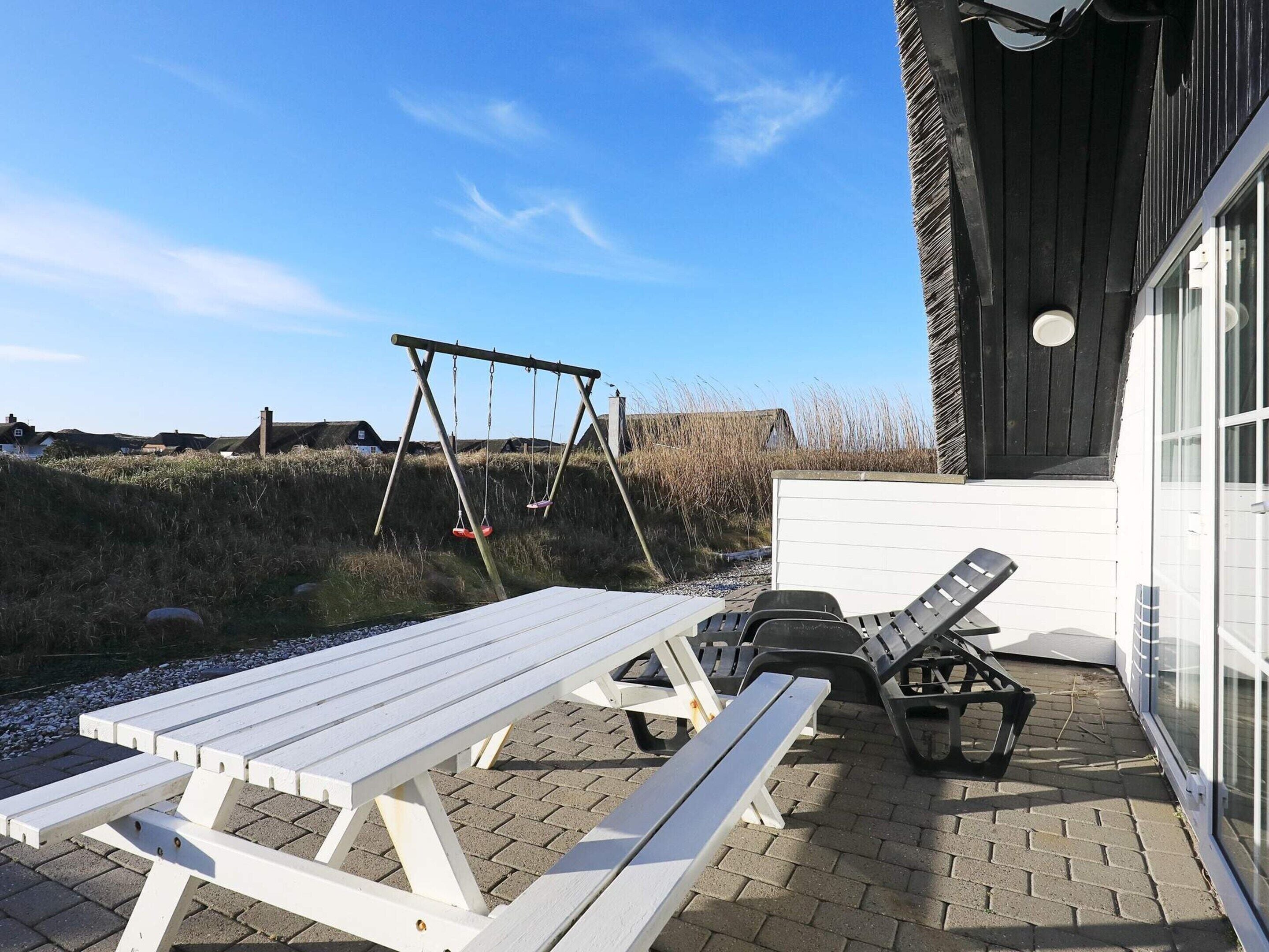 An outdoor seating area with picnic tables and lounge chairs, a swing set, and a building with a large window.