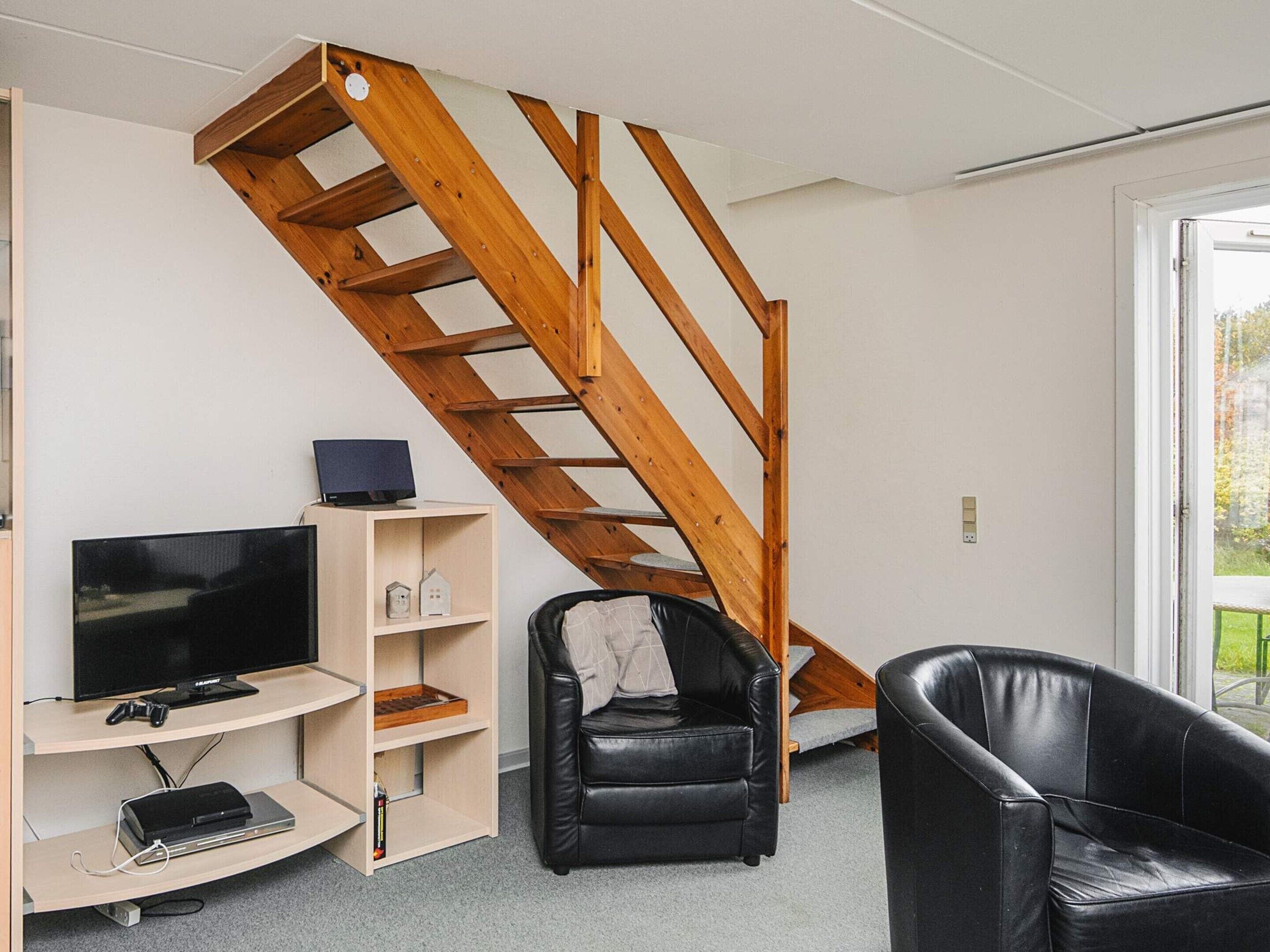Ein Zimmer mit einer Holztreppe, einem Fernseher, einer Spielkonsole und zwei schwarzen Ledersesseln.