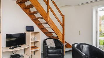 Ein Zimmer mit einer Holztreppe, einem Fernseher, einer Spielkonsole und zwei schwarzen Ledersesseln.