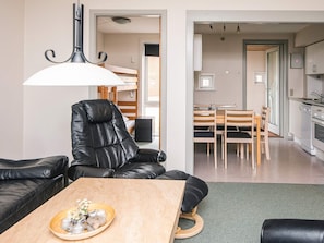 A modern living room with a leather sofa, a wooden coffee table with decorative items, a dining area with a wooden table and chairs, and a kitchen with white cabinets and a sink.