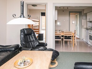 A modern living room with a leather sofa, a wooden coffee table with decorative items, a dining area with a wooden table and chairs, and a kitchen with white cabinets and a sink.