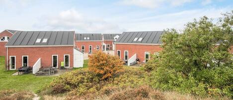 A row of red brick buildings with flat roofs and skylights, a grassy area with a bench, and a tree in the foreground.