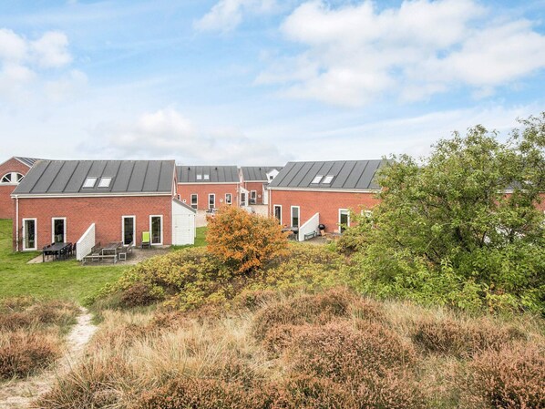 A row of red brick buildings with flat roofs and skylights, a grassy area with a bench, and a tree in the foreground.