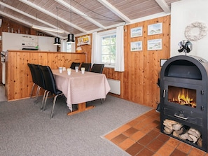 A dining area with a table set for four, a wood-paneled wall, a fireplace with logs, and a kitchen in the background. - Sun-kissed Holiday Home in BlÃ¥vand near Sea (Blavand)