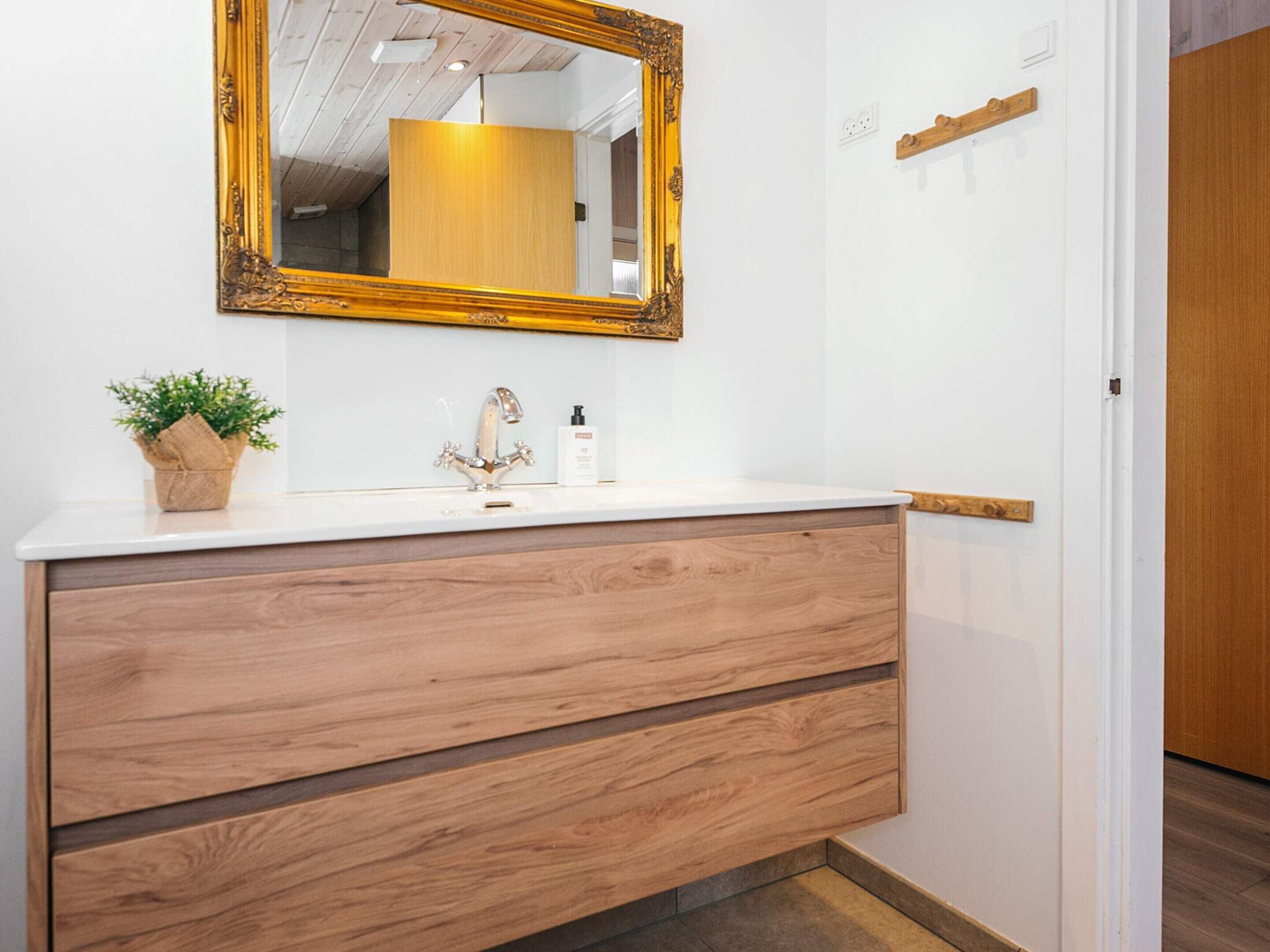 A bathroom with a white vanity, wooden drawers, a gold-framed mirror, a potted plant, and a bottle of hand soap.