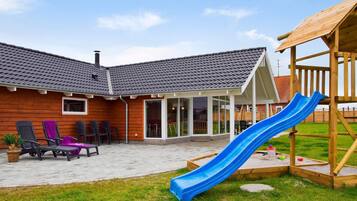 A backyard with a wooden playhouse, a slide, outdoor seating, and a covered patio area.