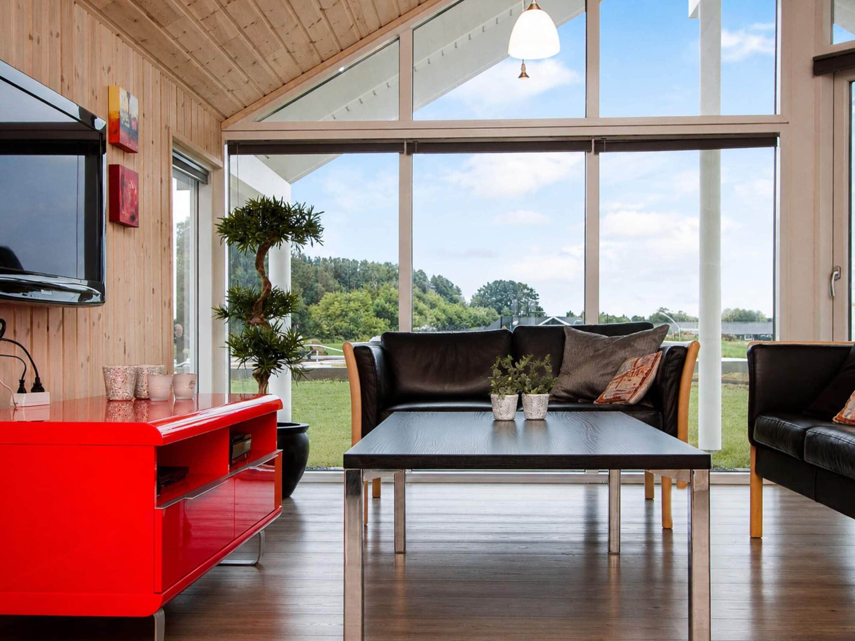 Ein modernes Wohnzimmer mit Holzboden, einer roten Unterhaltungseinheit, einem schwarzen Ledersofa, einem Couchtisch mit Glasplatte und einem großen Fenster mit Blick ins Freie.