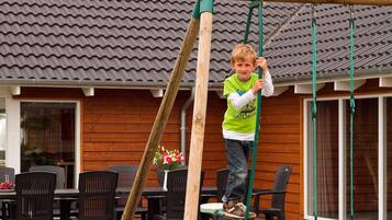 Un enfant joue sur une balançoire en bois dans un espace extérieur résidentiel, avec un toit en tuiles et des murs en bois.
