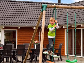 Un enfant joue sur une balançoire en bois dans un espace extérieur résidentiel, avec un toit en tuiles et des murs en bois.