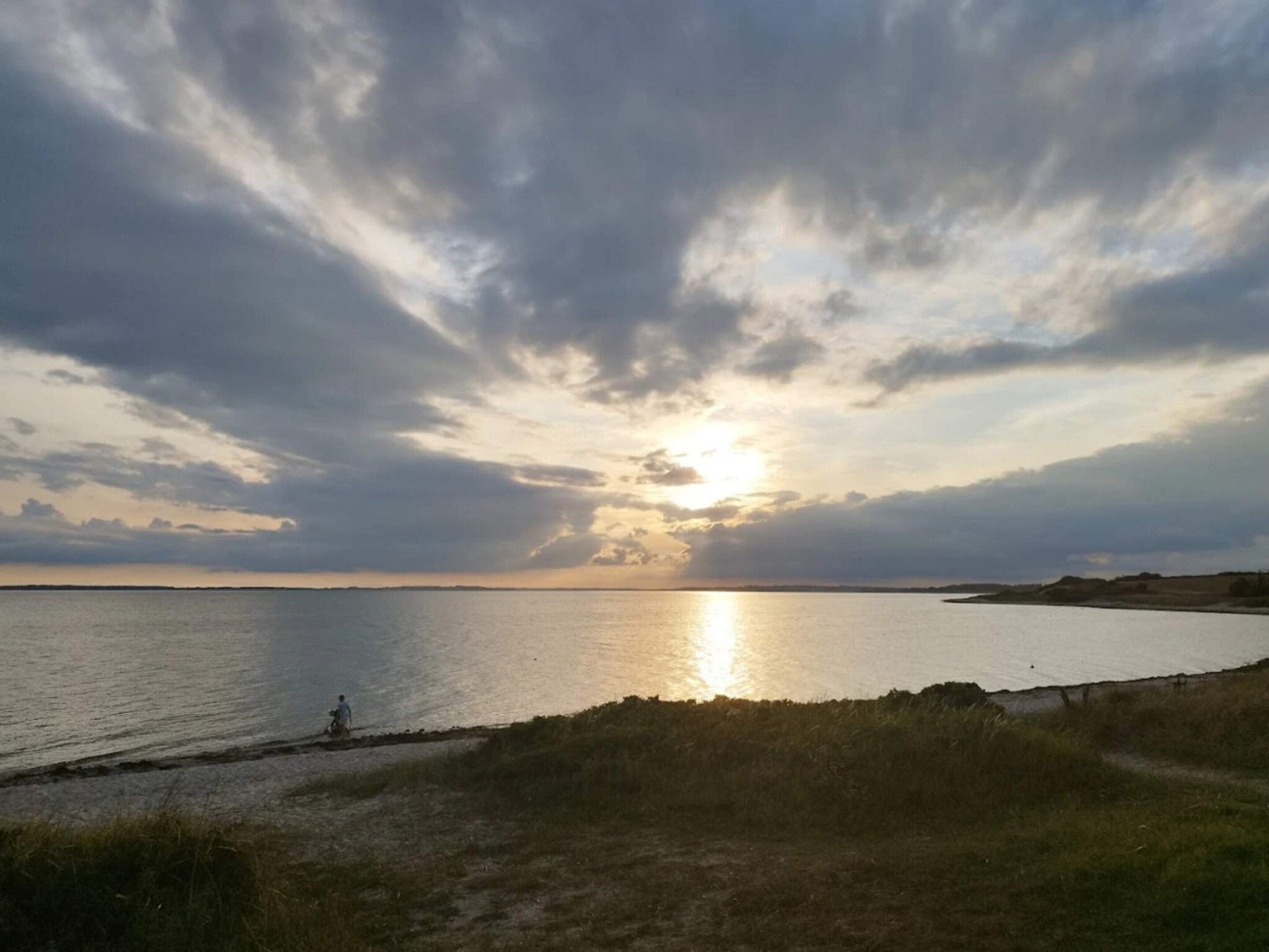 Un paysage côtier avec un coucher de soleil sur l’eau, une plage de sable et une personne qui se promène le long du rivage.