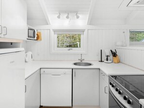 A compact kitchen with white cabinetry, a built-in dishwasher, a stove, and a window with a view of greenery. - 4 Star Holiday Home in Albaek (Aalbaek)