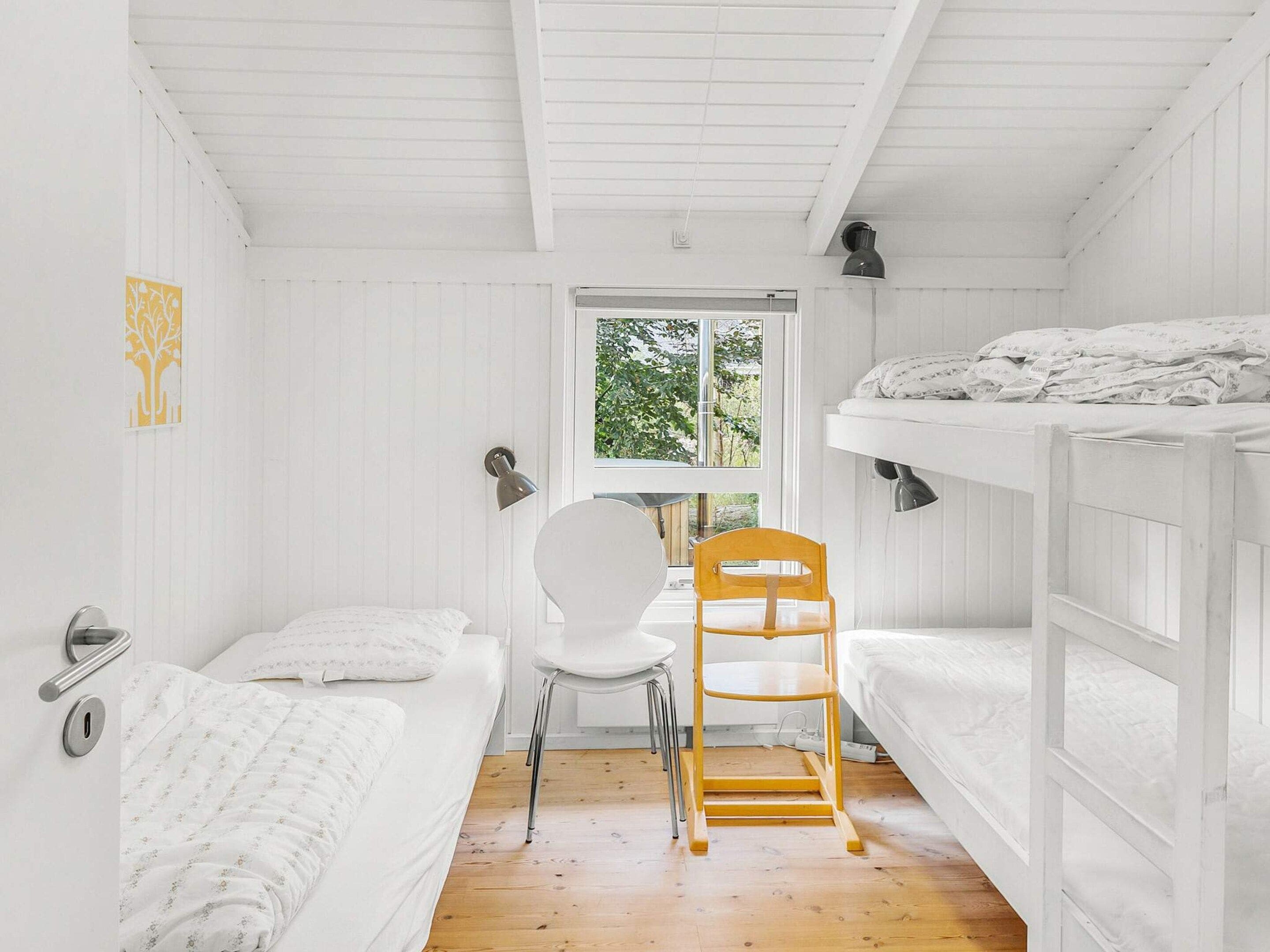 A small, white room with bunk beds, a single chair, and a window providing a view of greenery outside.