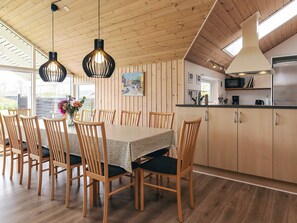 A dining area with a table and chairs, wooden walls, a kitchen with cabinets, and hanging pendant lights. - 4 Star Holiday Home in Idestrup (Idestrup)