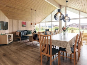 A spacious dining area with a long table and multiple chairs, a cozy living room with a sofa and a television, and large windows offering a view of the outdoors. - 4 Star Holiday Home in Idestrup (Idestrup)