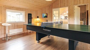 Habitació amb taula de bitlles, parets de fusta, una finestra i una porta que dona a una cuina.