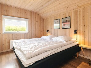 A bedroom with a wooden paneled wall, a large bed with white bedding, two wall-mounted lamps, and a window with a view of greenery. - 4 Star Holiday Home in Idestrup (Idestrup)
