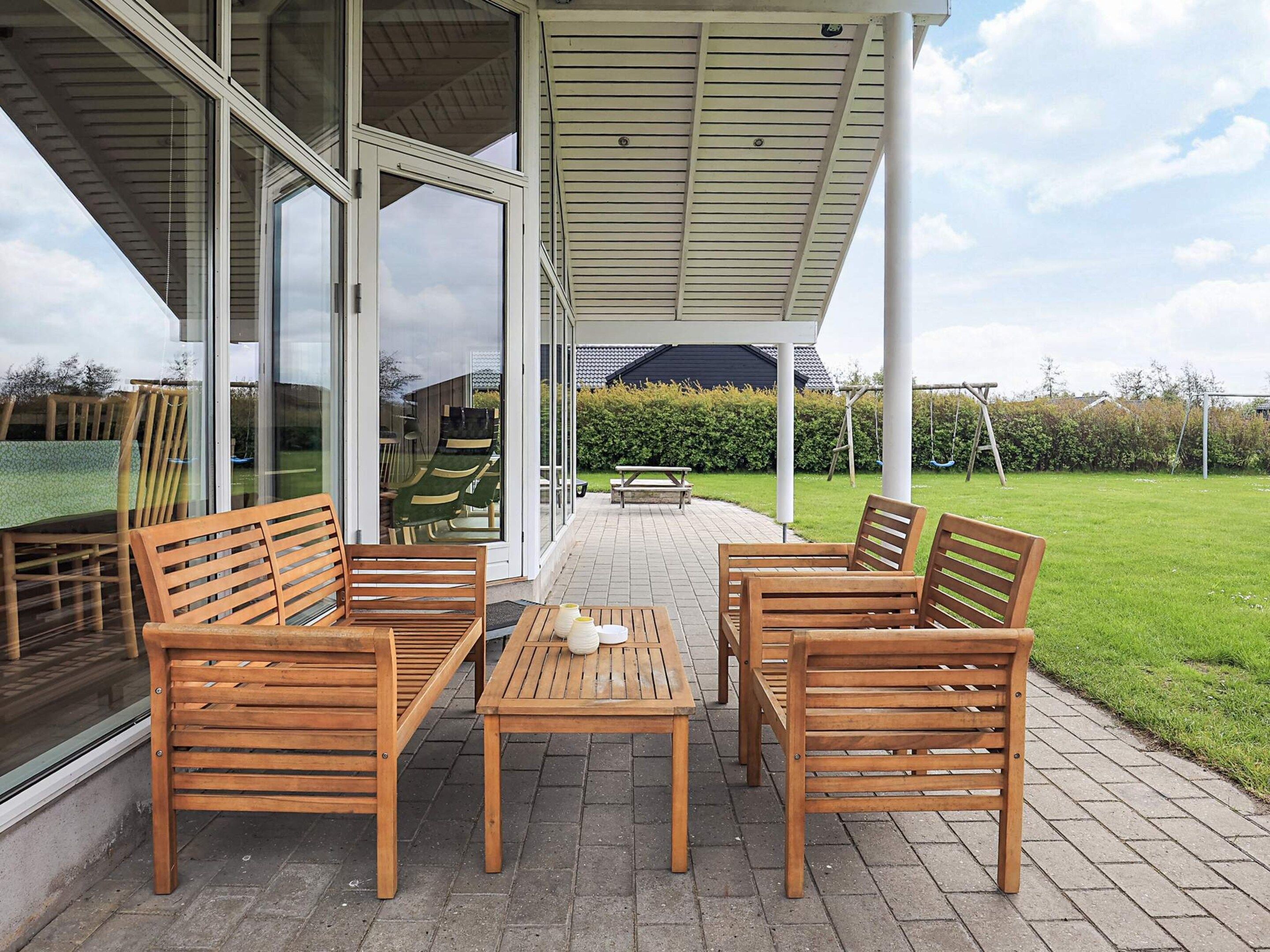 Une terrasse couverte avec des meubles en bois, une porte vitrée donnant sur un jardin et une vue dégagée sur les environs.