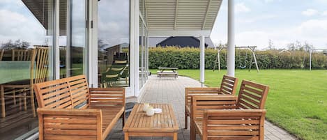 Un patio cubierto con mobiliario de madera, una puerta de vidrio que da a un jardín y una vista despejada del entorno.