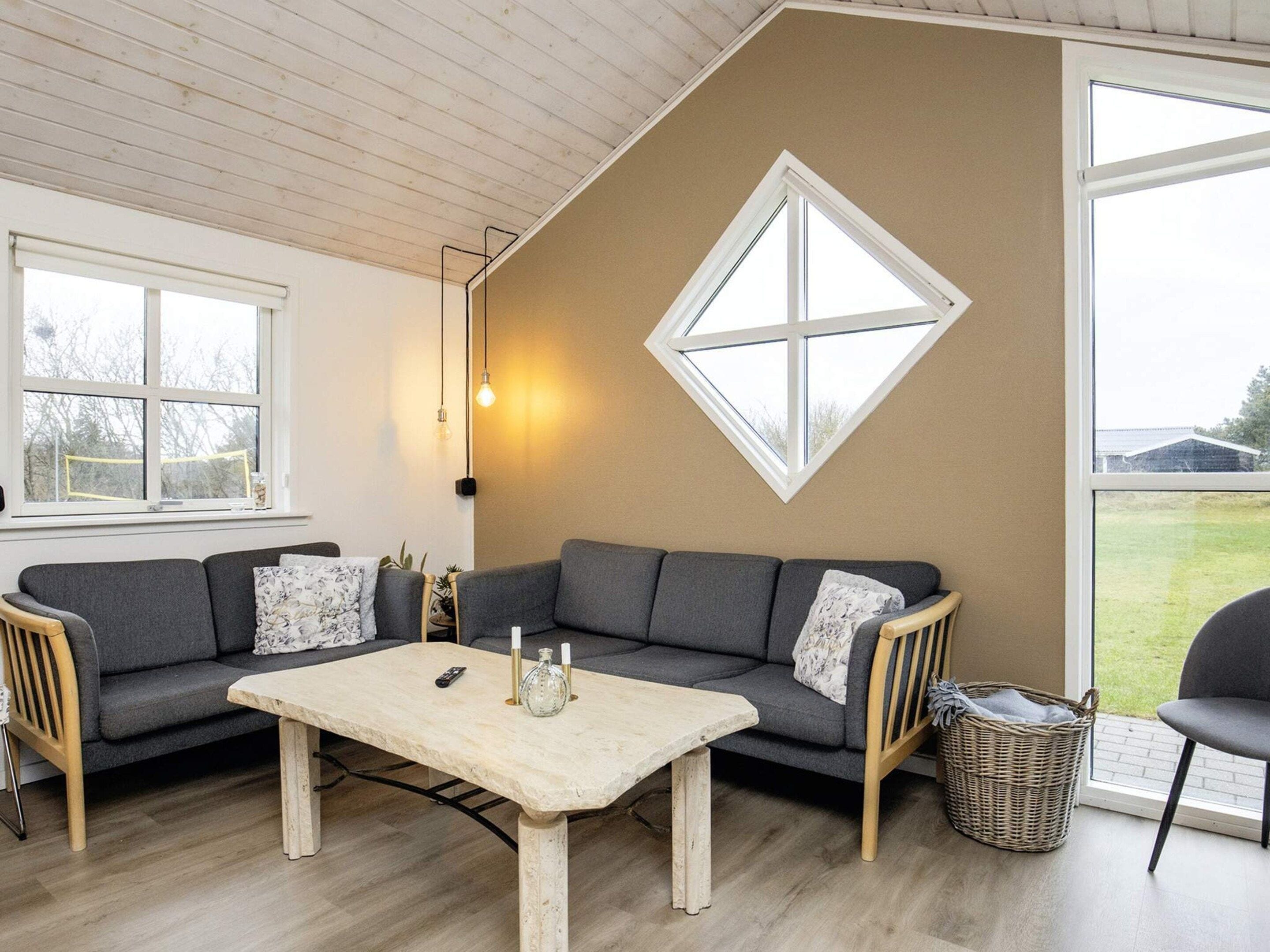 A cozy living room with a grey sofa, a wooden coffee table, and a large window with a diamond-shaped view.